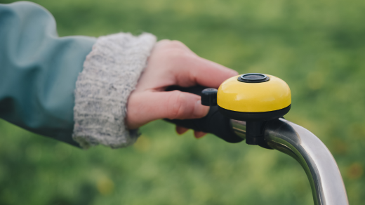 A person's hand rings a yellow bicycle bell. The bell is mounted on the bicycle handlebar.