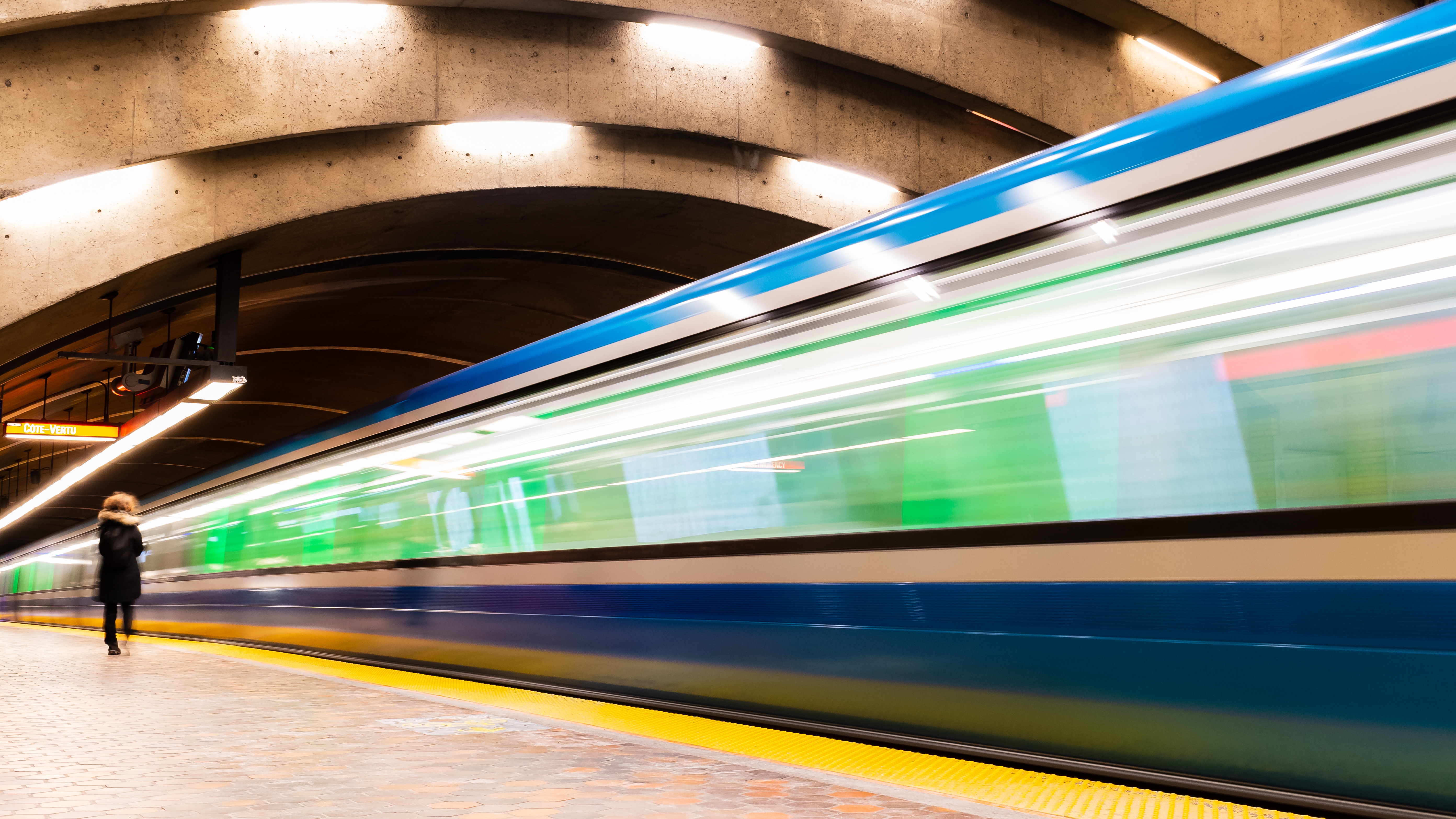 A train pulls into an empty indoor station. The image is out of focus and blurry, suggesting that the train is in motion as it approaches its stop. The station has a curved ceiling with lights, and one person walks along the platform.