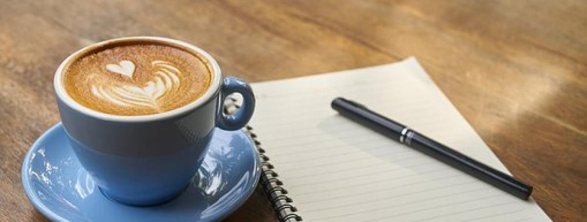 Une tasse de café avec un cœur dessiné dans le lait, sur un table en bois, à côté d'un cahier de note et d'un crayon.