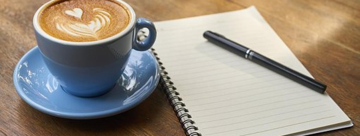 Une tasse de café latté avec un cœur formé dans la mousse, déposée sur une table en bois, à côté d'un pad de note.