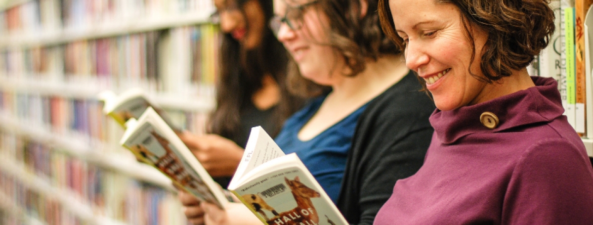 Three women standing in front of a row of bookshelves each reading a book.