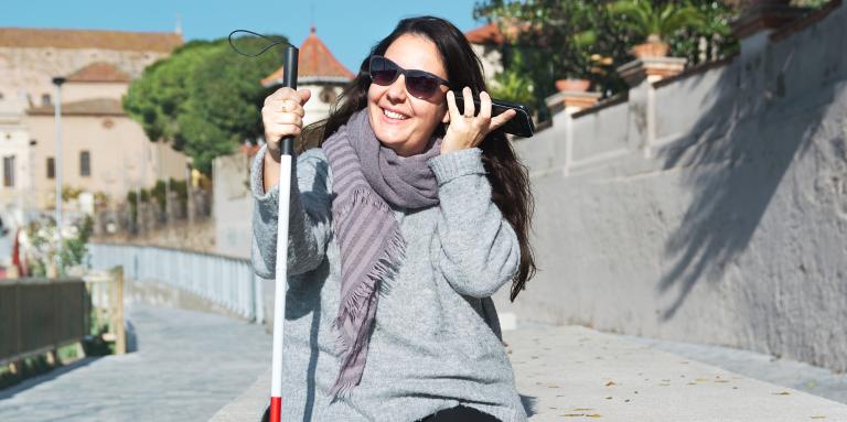 A young woman sits alone outdoors. In her left hand, she is holding her smartphone up to her ear. In her right hand, she holds her white cane