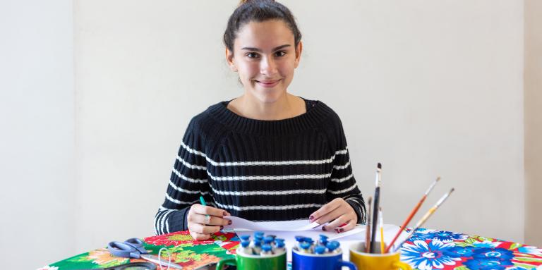 A young woman sits at a table making art with pencils, paper, scissors, magnifying glass, brush and paints in colourful mugs.