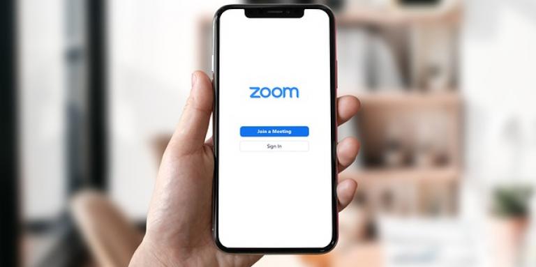 A hand holds up an iPhone 11. On the screen, the Zoom app is displayed.