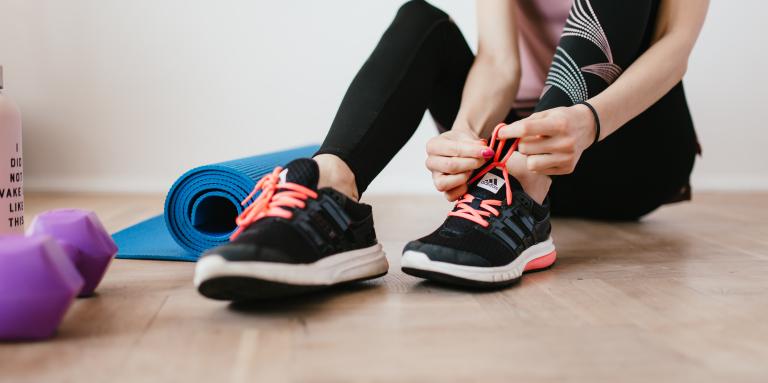A woman sitting down to tie up her running shoes. A yoga mat and dumbbell are beside her.