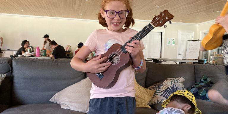 A young girl with red hair and glasses is standing while playing a ukelele in the Rec Hall. She has a big smile on her face.