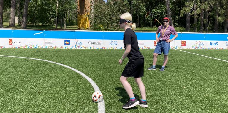Girl on the soccer pitch wearing a blindfold, about to kick the ball.