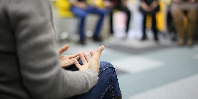 A person sits in a group circle. Their hands are clasped in their lap.