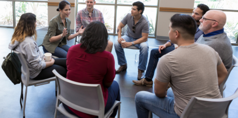 A support group of eight people sit in chairs in a circle and engage in lively conversation. 