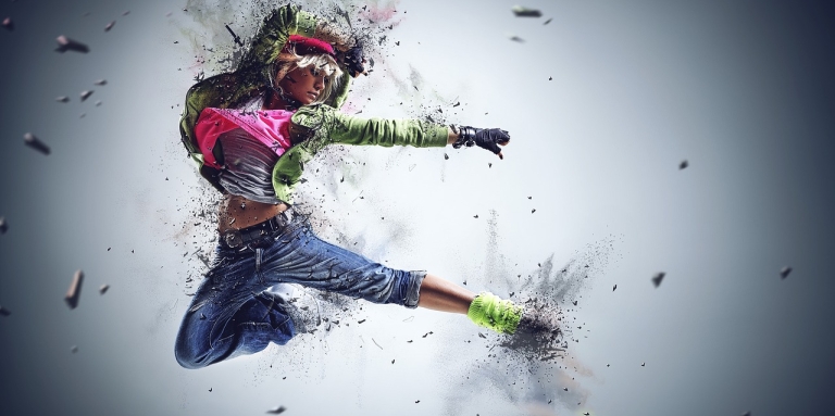 Image of young white woman jumping in the air in front of a silver screen. She is wearing cropped jeans, a white and pink shirt, a green jacket and green socks.