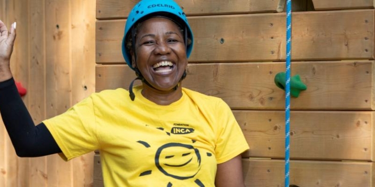 A black woman is standing in front of the climbing tower, smiling and waving with one hand. She is wearing a blue helmet and a yellow CNIB tshirt with a smiley face.