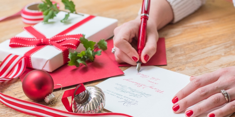 A woman is writing a message inside a card. There are gifts and ornaments and ribbons on the table.