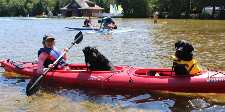 A woman is sitting in a kayak holding a paddle. There are two black guide dogs with her in the boat.