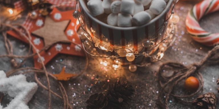 a mug filled with mini marshmallows is on a wooden table with candy canes, wooden ornaments, and tiny white lights.