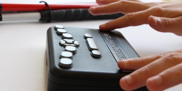 Hands reading the Orbit Braille Reader with a white cane in the background