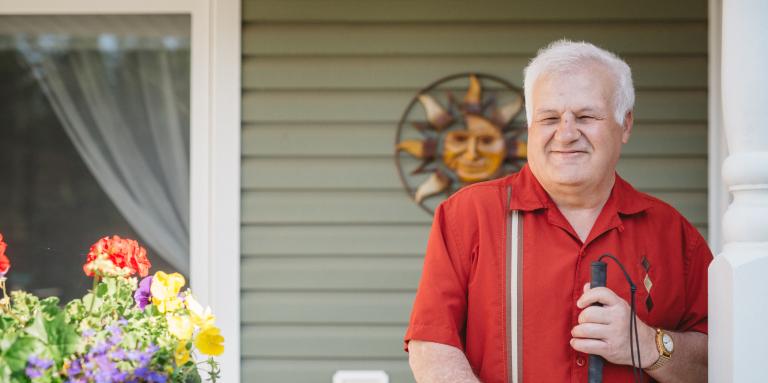Man with grey hair, wearing a red shirt, holding his white cane by his side, standing on his front porch.