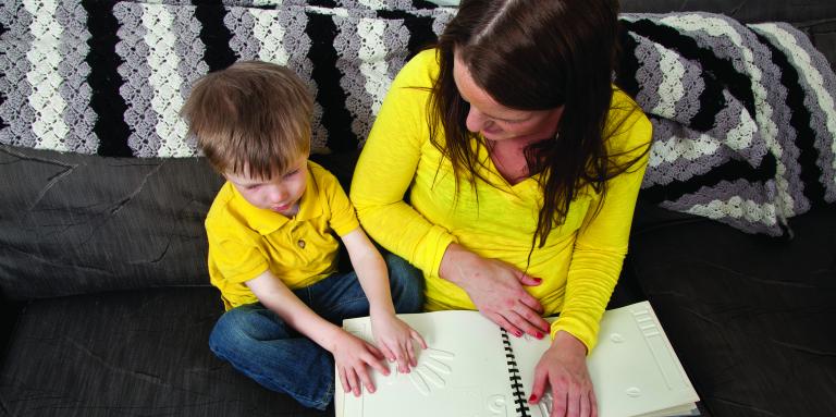 Un jeune garçon et sa mère, tous deux vêtus de chemises jaunes, parcourent un livre tactile assis sur un canapé.