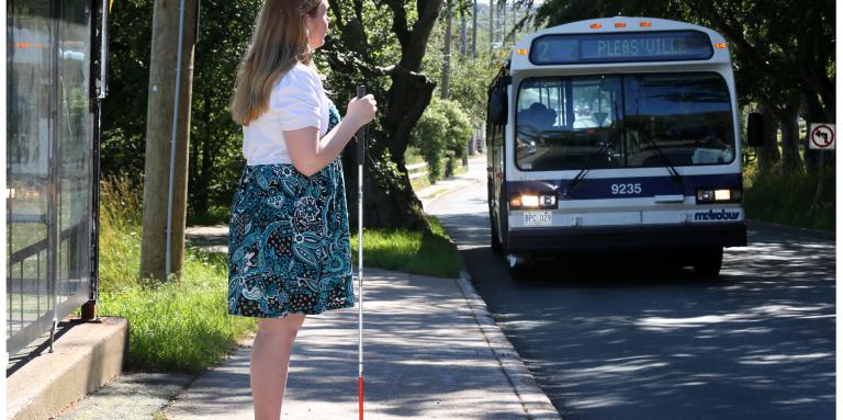 Young woman standing at a bus stop holding her white cane in front. The bus is approaching.