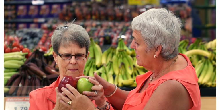 Two woman, wearing coral-coloured tops, are shopping for apples. 