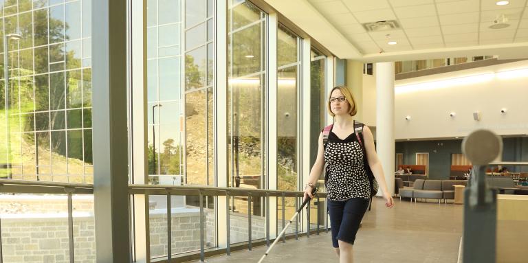 A young blond woman walking down a hallway with her white cane