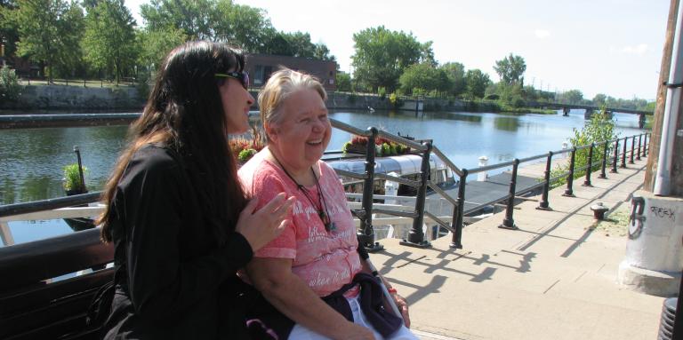 A blind woman sit on a bench outside next to a young lady, talking and laughing together