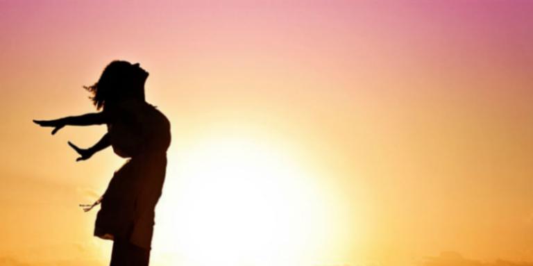 A woman stands silhouetted against a sunset, arms outstretched 