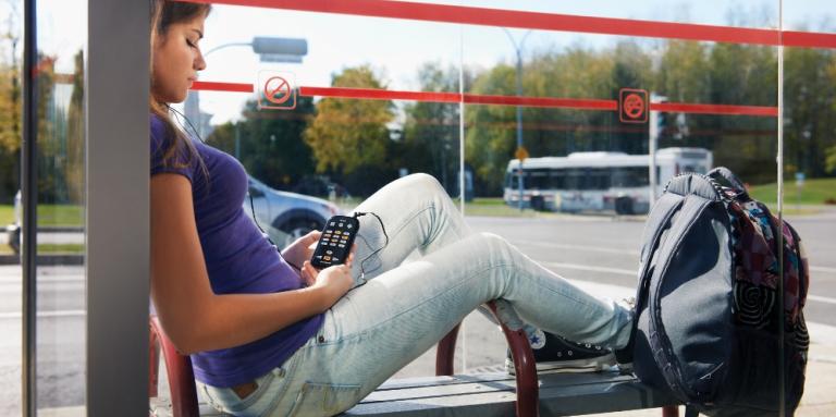 Photo of young woman relaxing, reading an audiobook, waiting at bus stop.