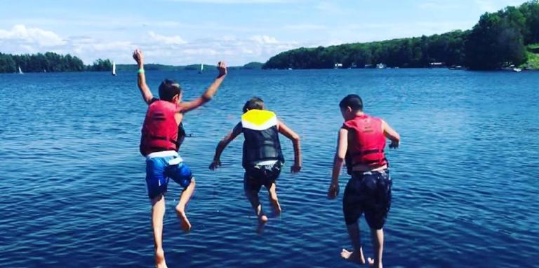 Three young boys wear life jackets and jump off the dock into the lake at CNIB Lake Joe.