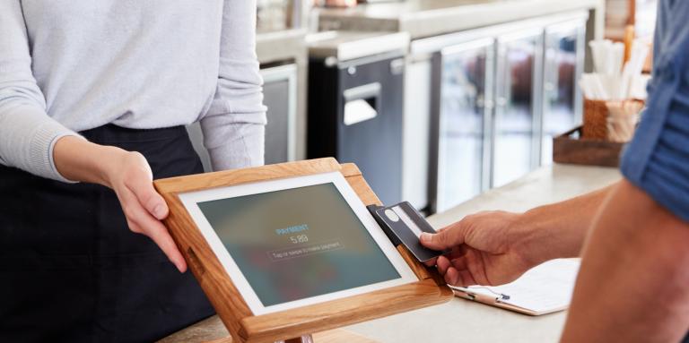 A person out of frame tapping their bank card on a touchscreen payment terminal, being presented by a café employee.