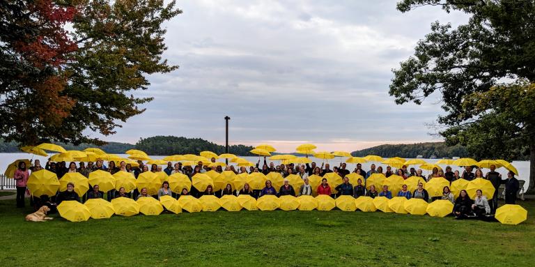 A large group of 50+ CNIB staff hold up yellow branded umbrellas on the shores of CNIB Lake Joe.