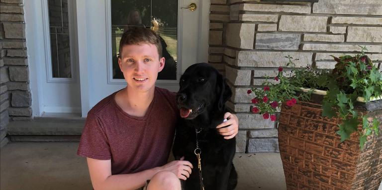 Jack McCormick sitting on the front step of house with his arm around his guide dog sitting at his side. 