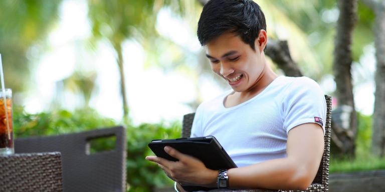 A man sits outside holding an iPad and smiles at what he sees on the screen.