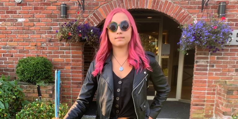 A cool teenager stands outside her house with her white cane in hand. She has pink hair and is wearing a leather jacket.