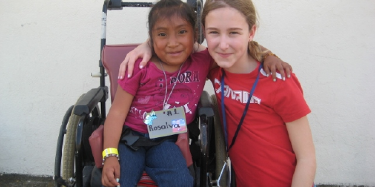 In Guatemala, a young girl crouches down next to a girl in a wheelchair. They have their arms around one another.