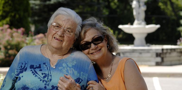 Two older women pose for a photo together. They are sitting outside near a water fountain.