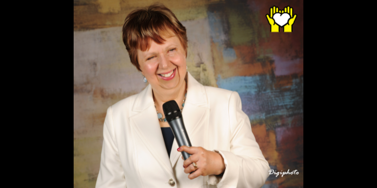 A photograph of Louise Burley smiling and holding a microphone in her left hand. A yellow graphic of hands holding a white heart outlined in black in the top right corner.