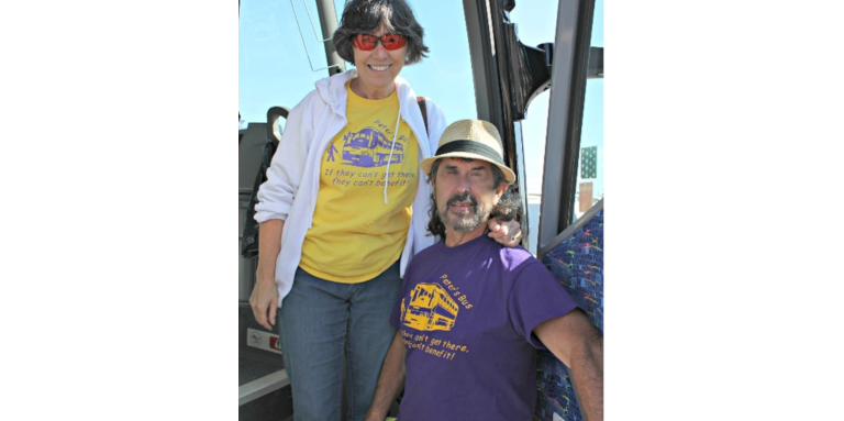 Peter and his wife Denise pose for a photograph at the bus entrance. Both are wearing t-shirts that say, "If they can't get there, they can't benefit."