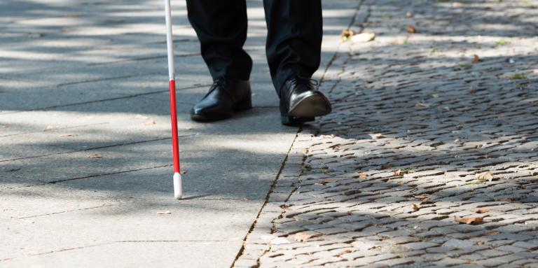  A white cane scans a sidewalk. In the background, mens shoes trail behind the tip of the cane.
