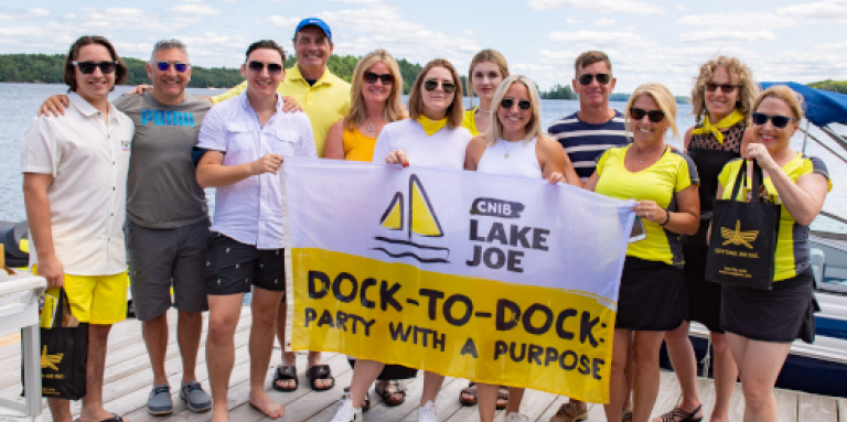 CNIB Muskoka Dock-to-Dock event volunteers posing at the CNIB Lake Joe boathouse, holding event flags.