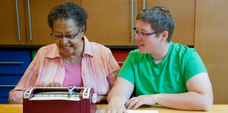 Deux femmes assises à une table sourient pendant que l'une d'elles utilise une machine braille.