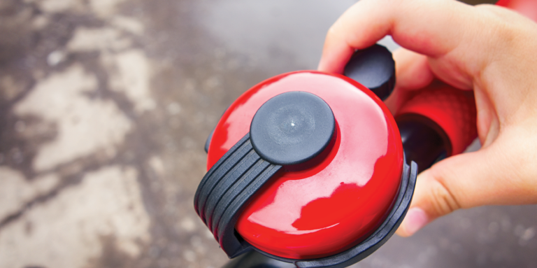 A person’s hand holds a red bicycle bell. The bell is mounted on the bicycle handlebar. 
