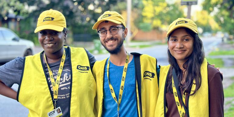 Three canvassers working on behalf of CNIB smile for the camera while standing on a sidewalk in a residential neighbourhood. Each canvasser is wearing a yellow CNIB baseball cap, vest, and lanyard with photo identification.