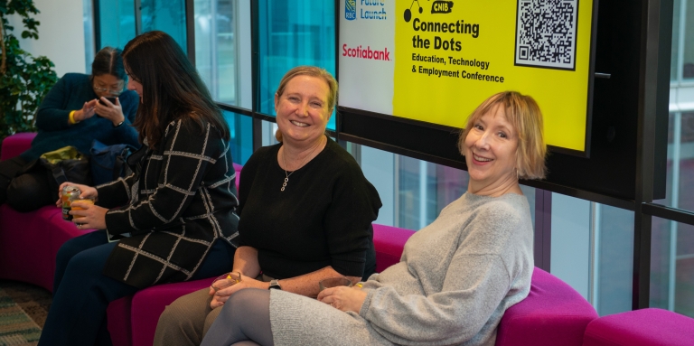 Two women sit on a couch with a Connecting the Dots sign behind them. 