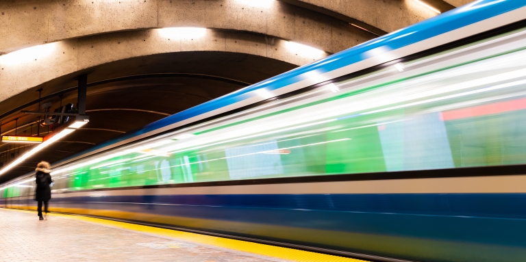 A train pulls into an empty indoor station. The image is out of focus and blurry, suggesting that the train is in motion as it approaches its stop. The station has a curved ceiling with lights, and one person walks along the platform.