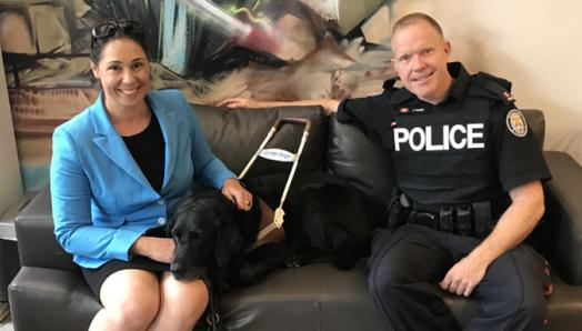 Victoria, her Guide Dog Alan, and a police officer sit on a couch.