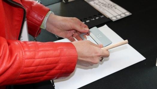 A woman holds a signature guide over a white page with a ballot and tactile ballot overlay beside her.