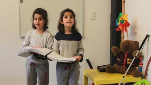 Two young children are holding on to the same braille book and walking around a classroom environment.
