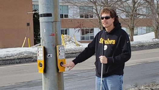 Bill Liggins se tient au coin de la rue à un panneau de signalisation piétonnière accessible. Il tient sa canne blanche dans la main gauche et appuie sur le bouton du passage pour piétons avec la main droite. 