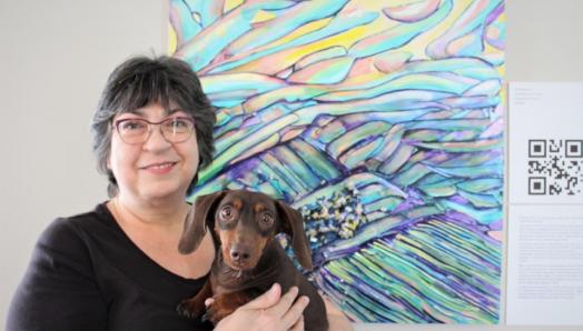 Robyn Rennie poses for a photo in front of her colourful artwork next to a digital printed QR Code. She is holding a small, chocolate-brown dog. 