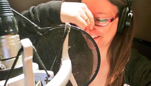 Dana est assise dans une cabine d'enregistrement et lit un livre. Elle se bouche le nez avec la main droite pour imiter la voix d'un personnage animé. Elle porte de gros écouteurs et parle dans un microphone. 